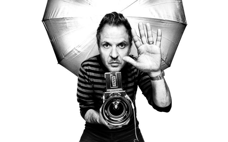 Platon standing in front of a photography umbrella, holding a camera, with his hand up