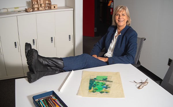 Kimberlee Douglas with her feet up at a table where she is drafting something on paper next to a box of colored pencils and a ruler