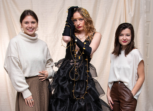 Lauren Schuler, Gina Zo, and Michaela Day standing together and smiling—Zo is wearing a formal, tiered, black gown with gold accessories—her look designed by Schuler and Day for the Grammys