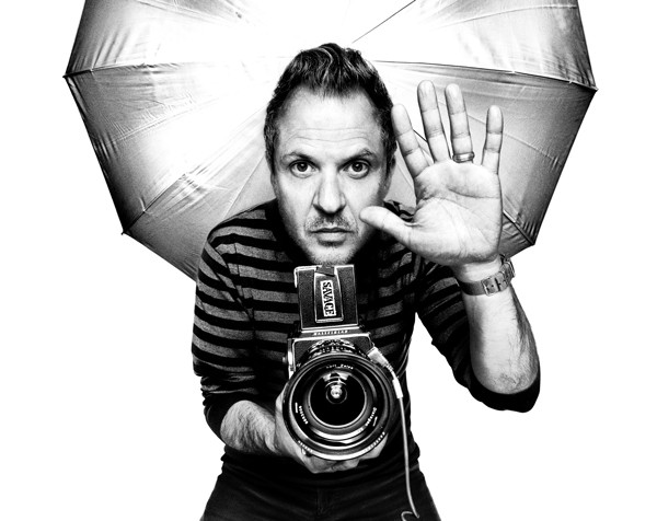 Platon standing in front of a photography umbrella, holding a camera, with his hand up